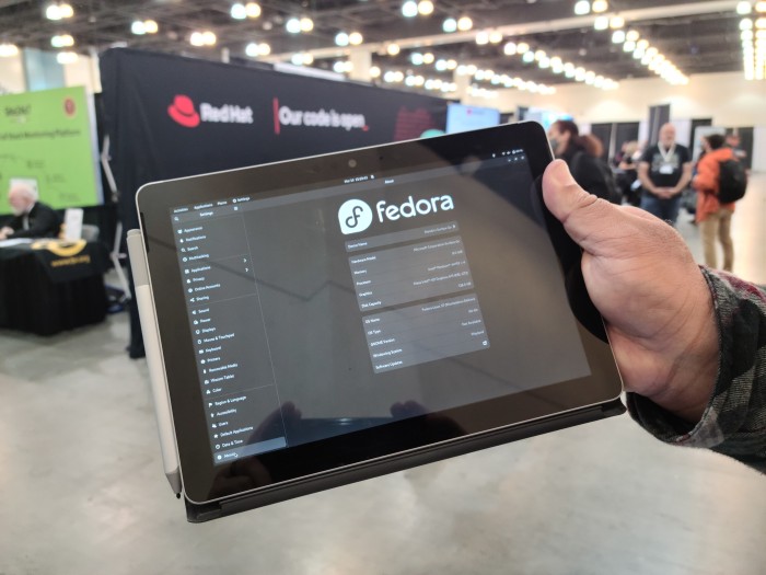A hand holding a Surface Go tablet which is displaying the GNOME Settings "About" page. It is running Fedora Linux 37.