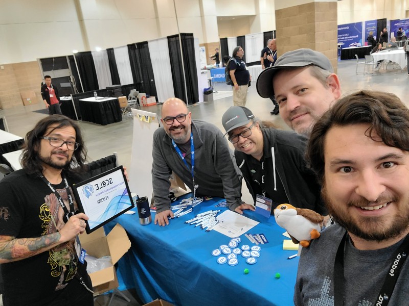 An elevated picture of five people standing around a blue-clothed conference expo table covered in pens, stickers, and other swag.